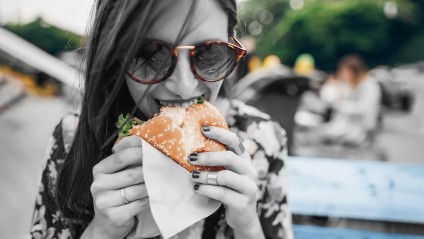 A young female with glasses eating a hamburger on an outside bench