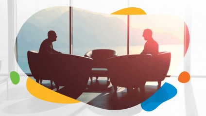 Two men sitting in chairs having a discussion in front of large windows
