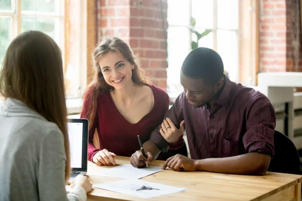A man signing home purchase agreement and his smiling wife sitting next and looking at the female real estate agent