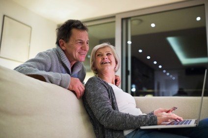 Beautiful senior couple at home in their living room, woman with laptop sitting on couch shopping online