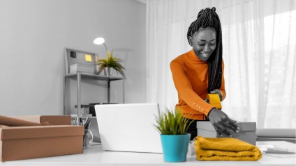 A young female in an orange sweater taping up a box next to a laptop