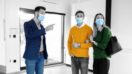 Young couple with masks and brightly colored clothes touring a home with their masked real estate agent.