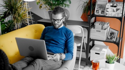 A male in a blue shirt laying on a yellow couch with his laptop