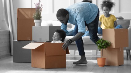 man wearing a blue shirt with polka dots and glasses pushing his child across the wooden floor in a moving box with his family watching behind him