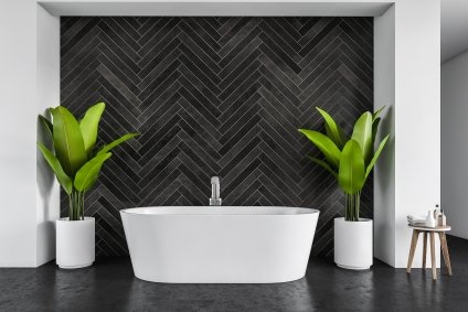 White bath against black tile wall in a big bathroom with plants in pots on the sides.