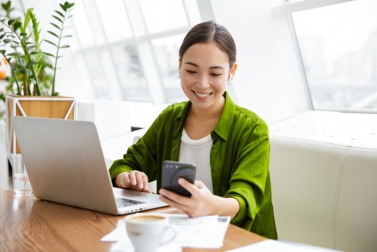 Woman in green shirt looking at phone while working at laptop