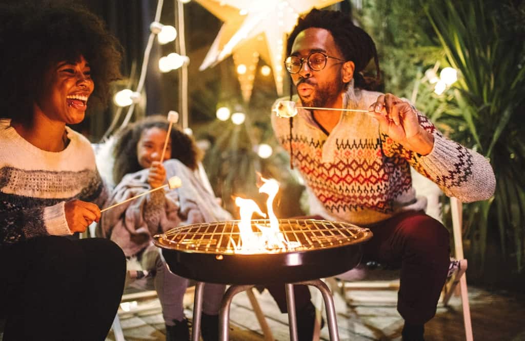 family surrounding a campfire father blowing on marshmallow while backyard camping