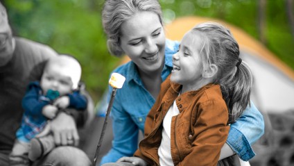 blonde mom wearing denim button up roasting marshmallows and holding her blonde daughter with her family in the background