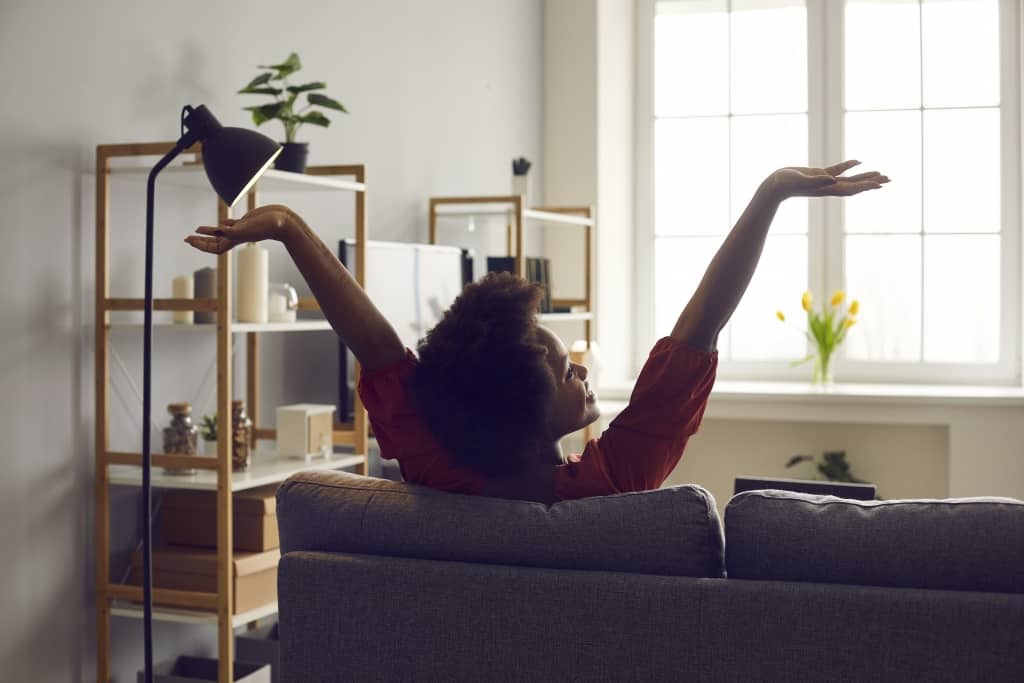 Back view of happy young woman stretching her arms sitting on couch in living-room