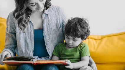 young mom wearing a stripped blue button down shirt smiling and helper her toddler son read a book in his book nook