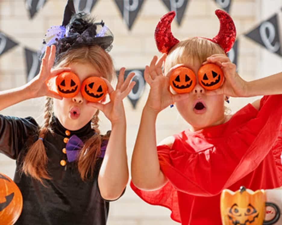 Two children wearing witch and devil halloween costumes, holding mini pumpkins to their eyes
