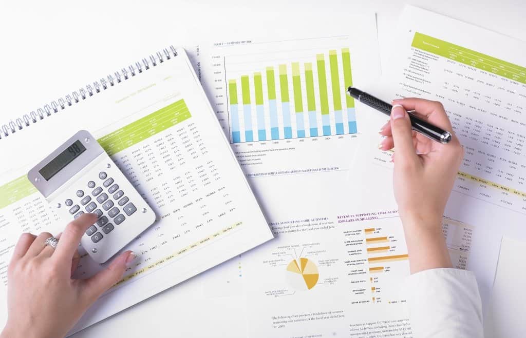 a woman\'s hand holding a pen and calculator with printed spreadsheets and graphs on a desk