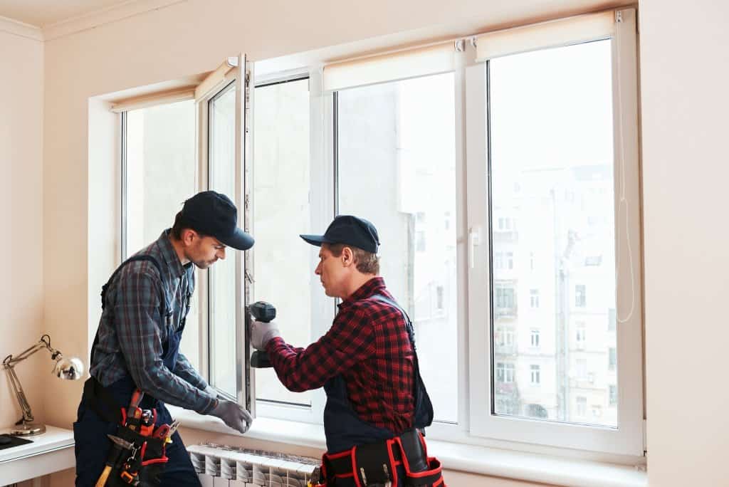 Two men wearing tool-belts working on a window casing with a power tool