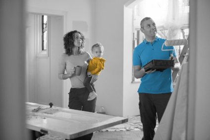 Man in a blue shirt holds a paint roller while a woman holding a baby looks on